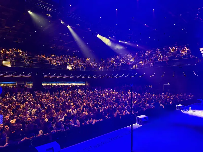 View from the side of the stage across a packed standing crowd and balcony under blue stage lighting