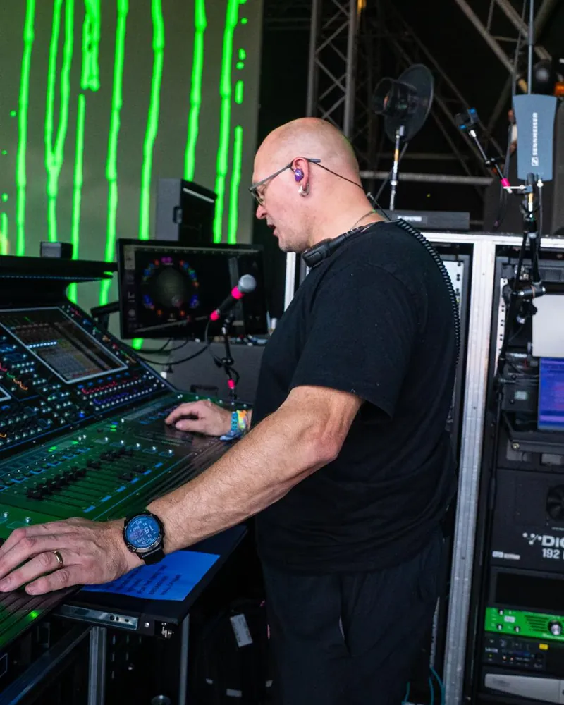 Mike Osman mixing at a DiGiCo console in front of a green video screen, wearing in-ear monitors, with racks and a Sennheiser antenna beside him